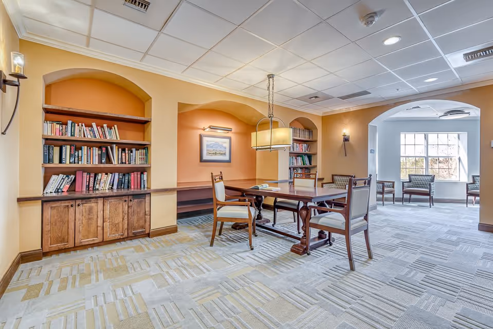 A well-lit interior room with built-in wooden bookshelves filled with books, a wooden table with four upholstered chairs, and a hanging light fixture above the table. The room has warm yellow and orange walls with arched alcoves and a patterned carpet. In the background, there is a seating area with several chairs near large windows letting in natural light.