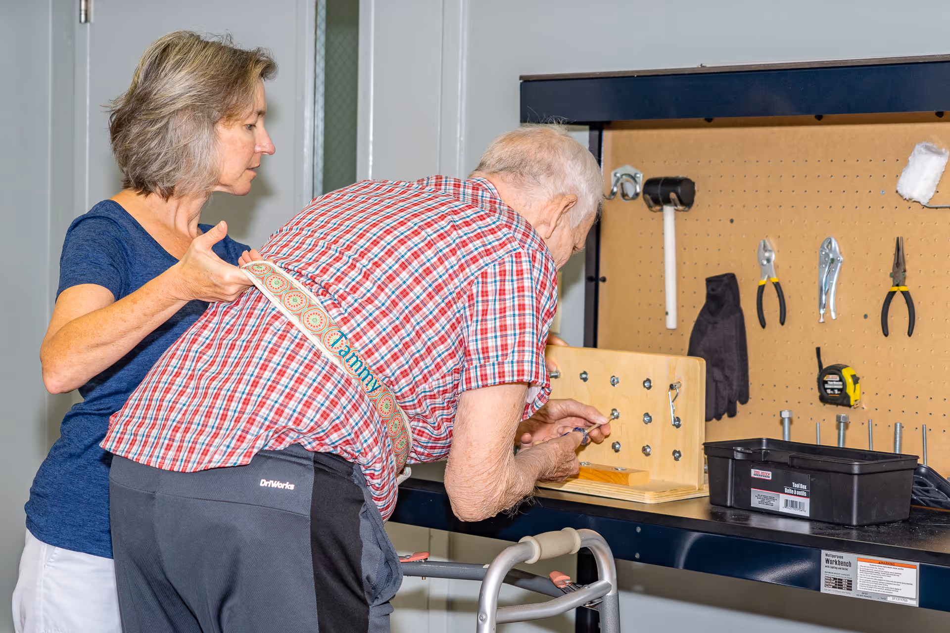 An elderly man using a walker is engaged in a hands-on activity at a workbench with tools, assisted by a woman standing beside him providing support.