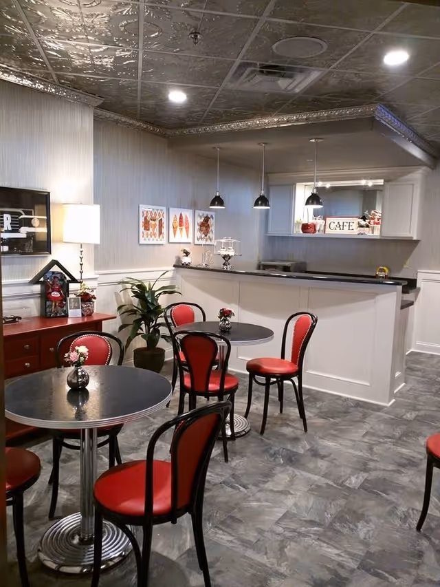 Interior view of a small cafe area with round tables and red cushioned chairs. The space features a white counter with three hanging pendant lights above it. There are decorative pictures on the wall, a potted plant, and a lamp on a red sideboard. The floor has a gray tile pattern and the ceiling has an ornate metallic design.