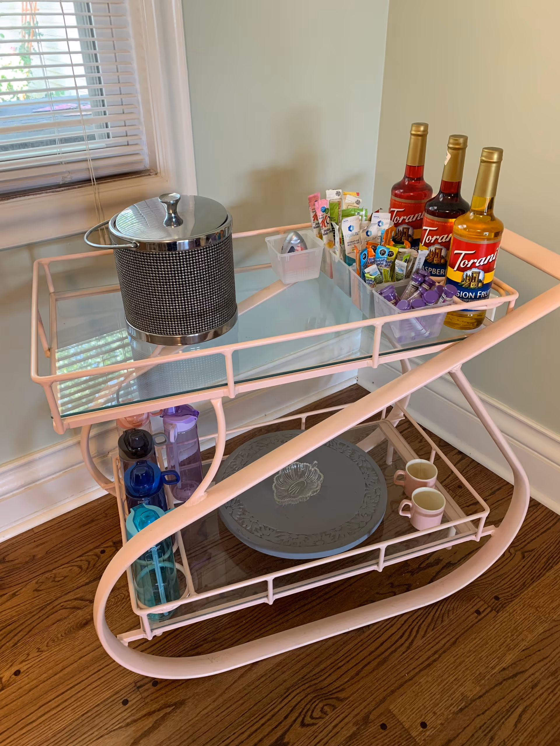 A pink metal and glass beverage cart with various drink supplies including three bottles of Torani syrup, an ice bucket, assorted drink mixers, water bottles, a decorative plate, and two pink mugs. The cart is positioned on a wooden floor near a window with white blinds.