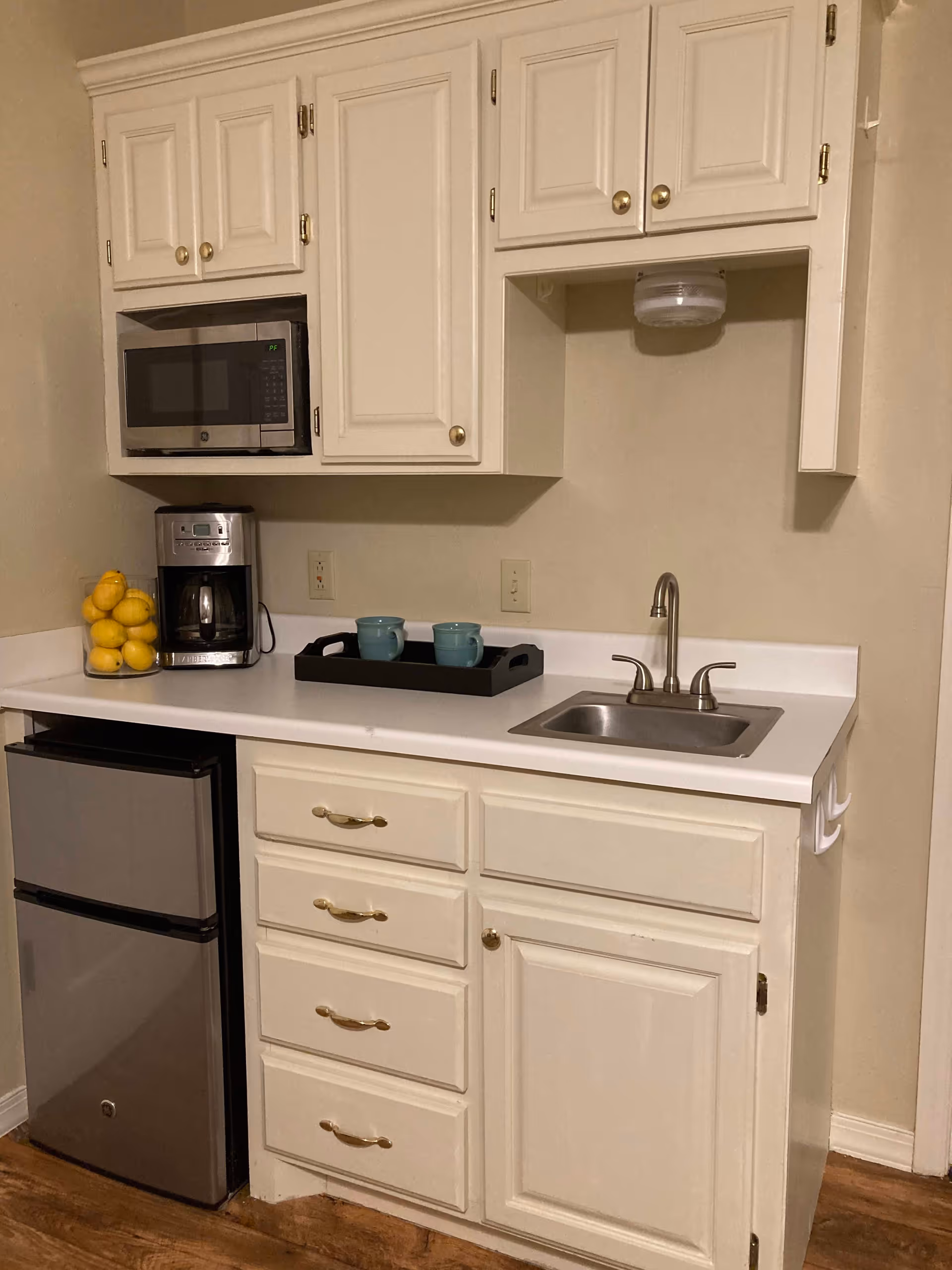 A small kitchen area with white cabinets and drawers, a stainless steel mini refrigerator, a coffee maker, a microwave, a sink with a faucet, and a tray holding two blue mugs. A glass container filled with lemons is also on the countertop.