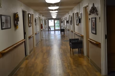 A long, well-lit hallway in a nursing and rehabilitation facility with wooden flooring, handrails on both sides, framed pictures and decorations on the walls, and a few chairs placed along the corridor. The hallway leads to a glass door at the far end.
