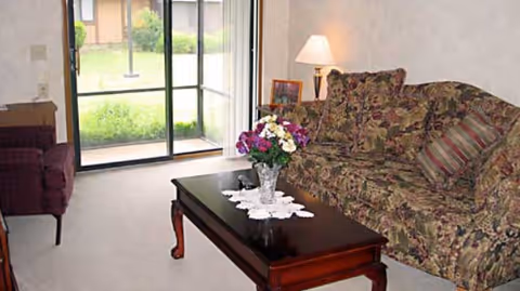 A cozy living room with a floral patterned sofa adorned with matching cushions, a wooden coffee table with a lace doily and a vase of colorful flowers, a side table with a lamp and a framed photo, and a sliding glass door leading to a green outdoor area.