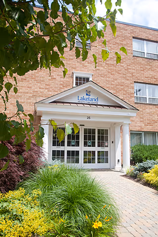 Entrance of Lakeland Health Care Center, a brick building with white columns and a glass door, surrounded by green plants and flowers along a paved walkway.
