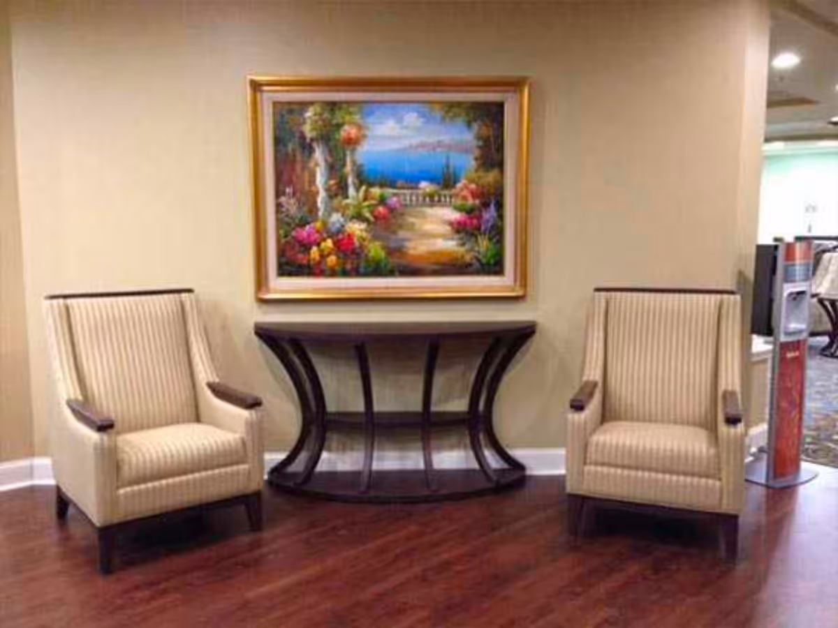 Two beige striped armchairs with dark wooden armrests are positioned on either side of a dark wooden console table with curved legs. Above the table hangs a colorful framed painting depicting a garden path leading to a scenic view of the sea with flowers and greenery.