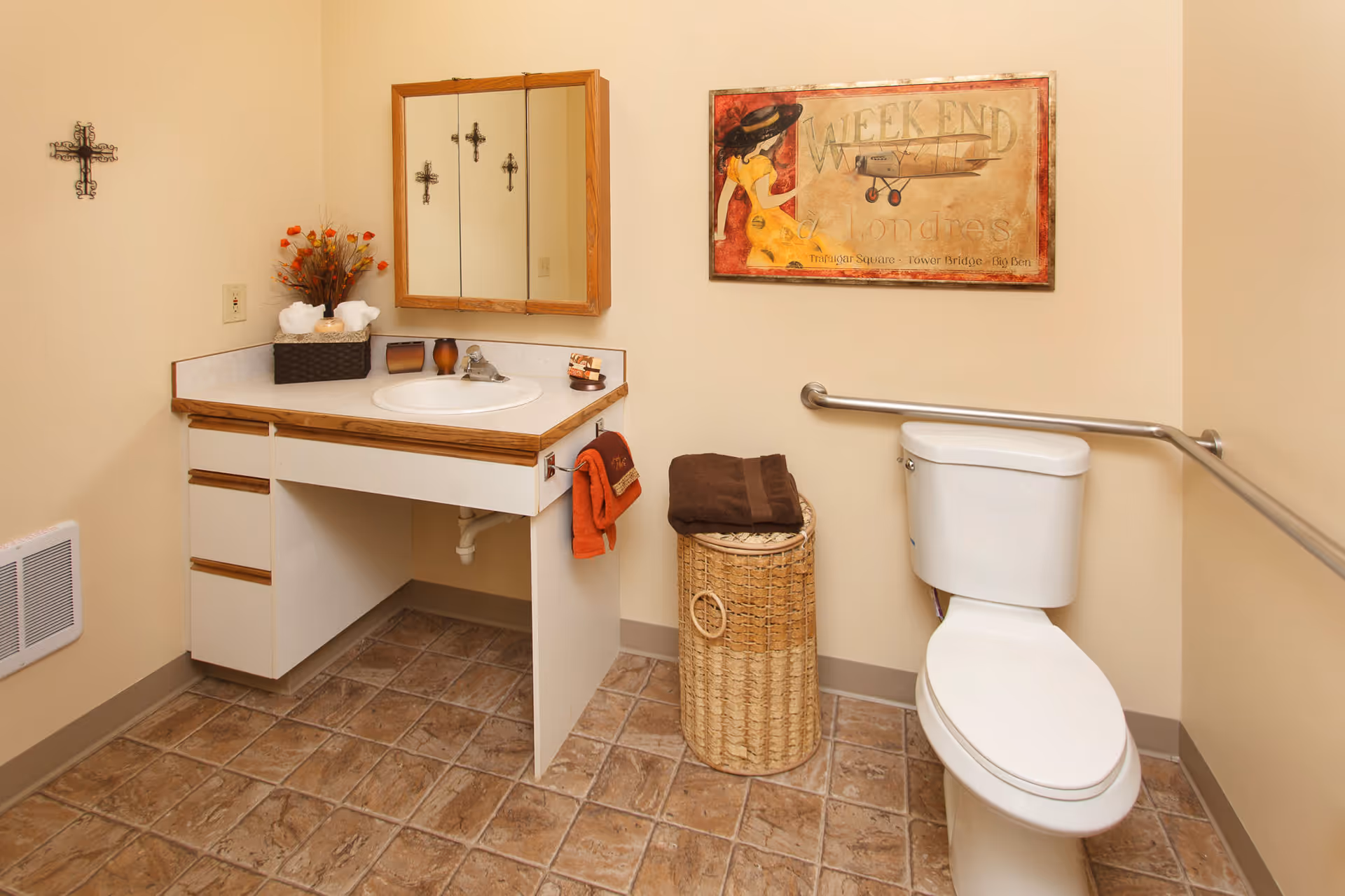 A bathroom with a white toilet equipped with a safety grab bar, a vanity with a sink and a mirror above it, a wicker laundry basket with folded towels on top, a wall decoration featuring a woman and an airplane, and a small cross hanging on the wall.