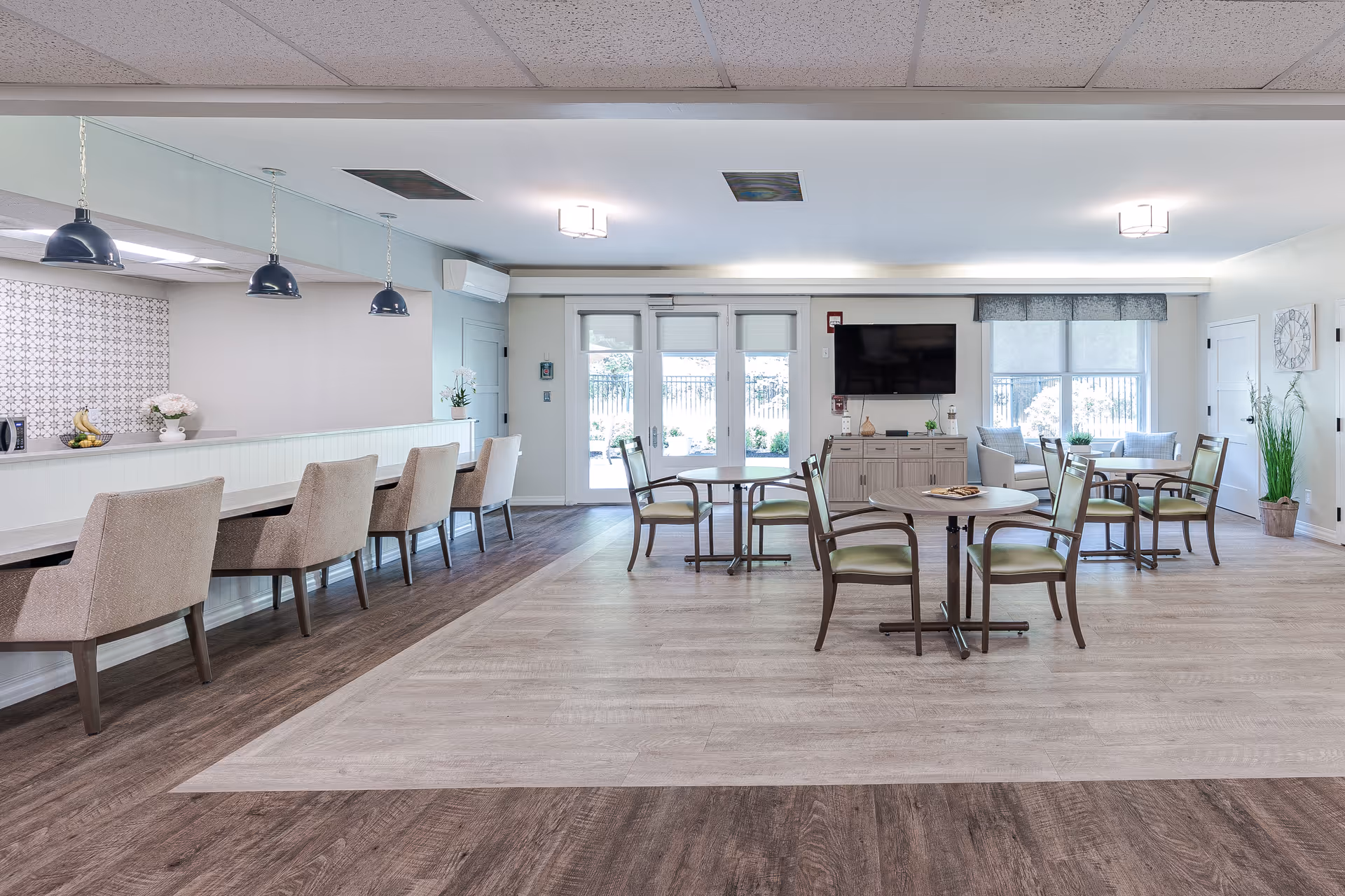 A bright and spacious common area with a combination of seating options including a long counter with upholstered chairs and several round tables with wooden chairs. The room features light-colored walls, large windows and glass doors letting in natural light, a mounted flat-screen TV, and decorative plants. The flooring is a mix of light and dark wood tones, and the ceiling has recessed lighting and hanging pendant lights above the counter.