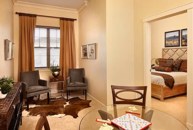 Interior of a senior living suite with a small sitting area of two chairs, a glass dining table in the foreground, and a bedroom visible through a doorway.