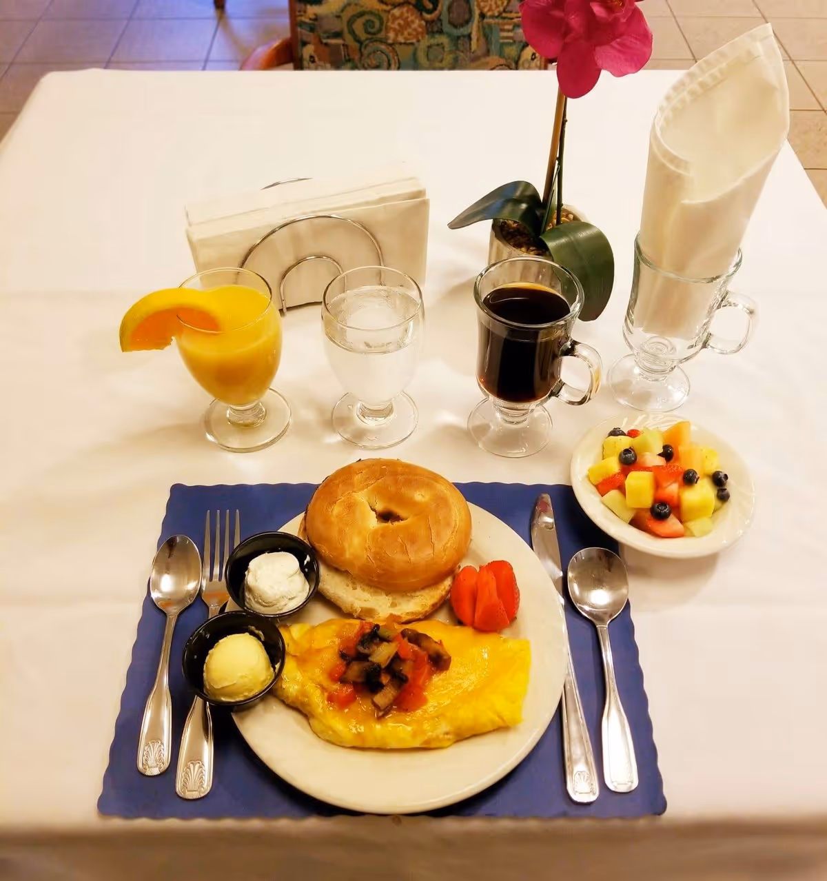 A breakfast meal set on a white tablecloth with a blue placemat. The meal includes a folded omelette topped with diced vegetables, a bagel with cream cheese, a small serving of sliced tomatoes, and a bowl of mixed fruit with blueberries, melon, and watermelon. There are three beverages: orange juice with an orange slice, a glass of water, and a cup of black coffee. Silverware includes a fork, knife, and two spoons. A napkin holder with white napkins and a glass holding a folded white cloth napkin are also on the table, along with a small potted orchid plant.
