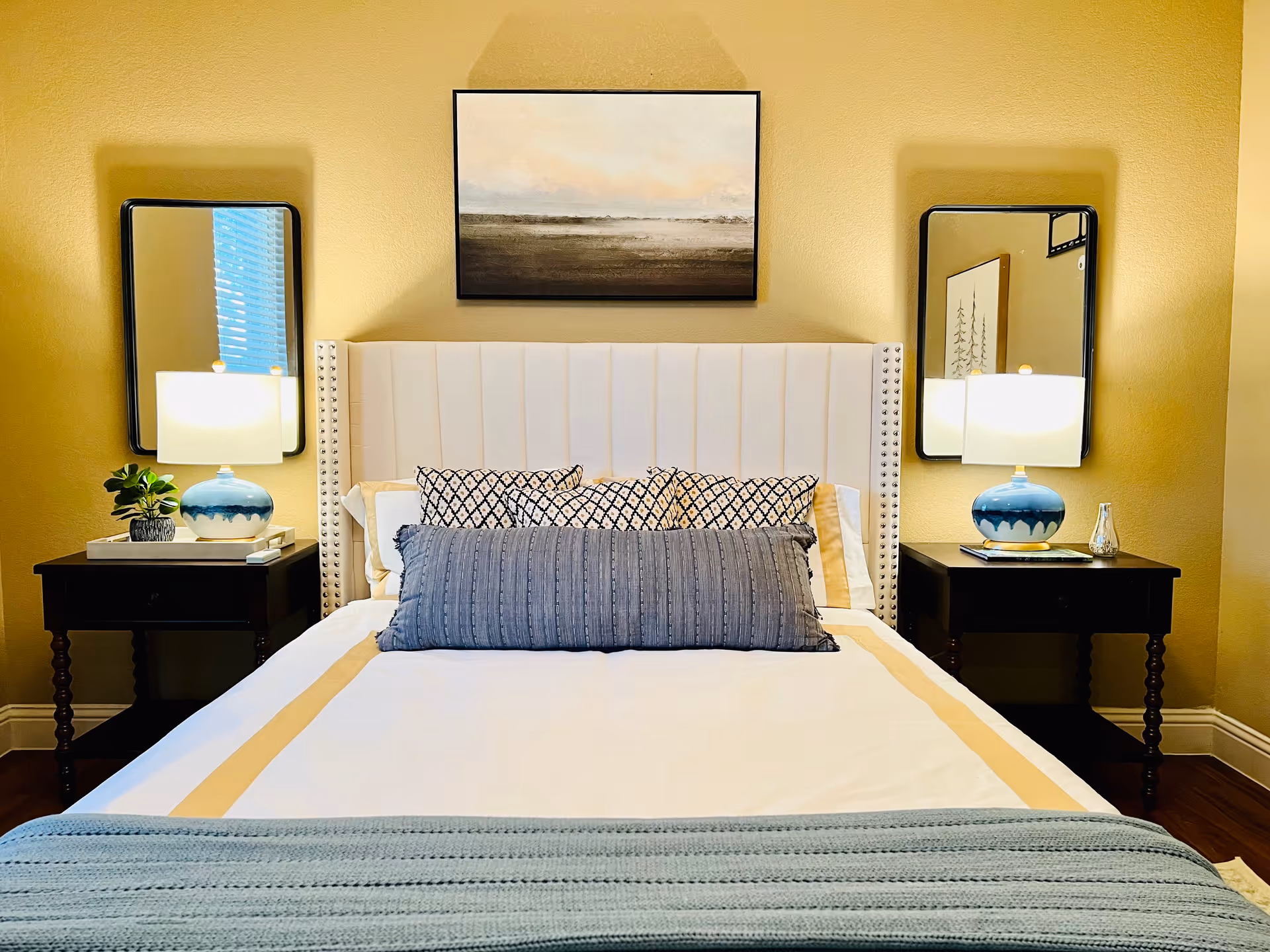Neatly made bed with an upholstered headboard, decorative pillows, bedside tables with lamps and mirrors, and artwork above.