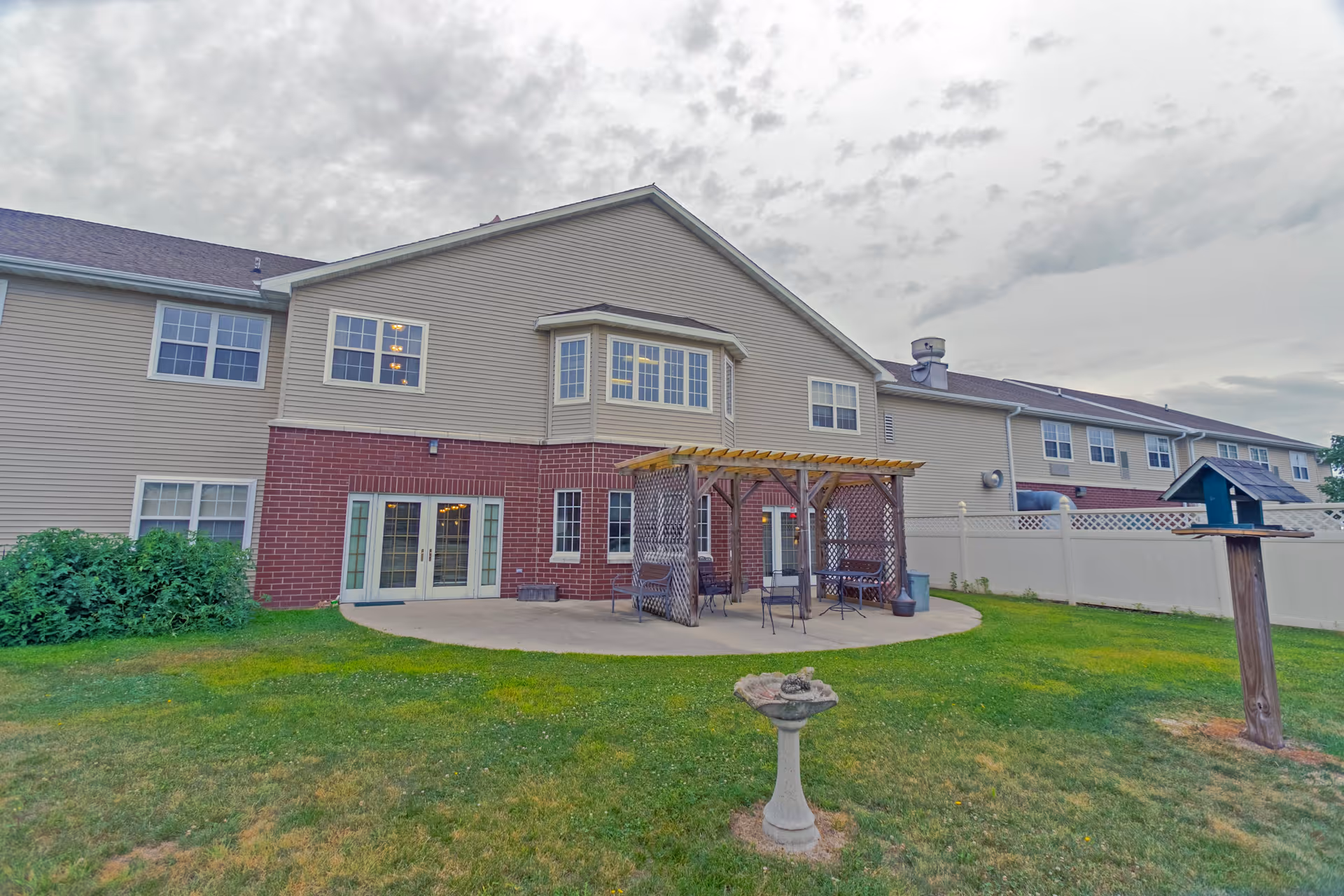 Exterior view of a two-story senior living facility building with beige siding and red brick accents. The building features multiple windows and a patio area with a wooden pergola, outdoor seating, and a bird feeder on a post in the grassy yard.