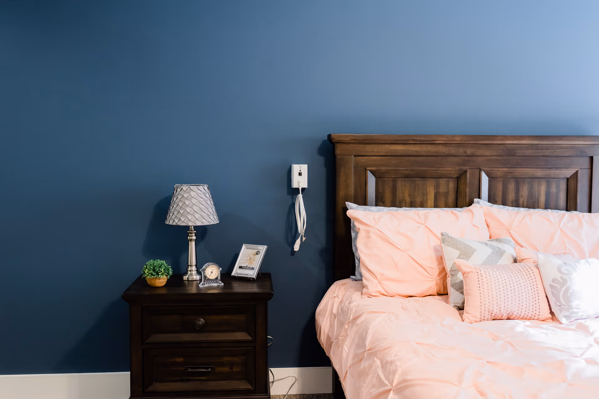 A bedroom corner featuring a dark wooden headboard and a bed with light pink bedding and multiple pillows. Next to the bed is a dark wooden nightstand with a small green plant, a silver lamp with a patterned shade, a small clock, and a framed photo. The wall behind is painted dark blue with a white baseboard and a white emergency call button mounted on the wall.