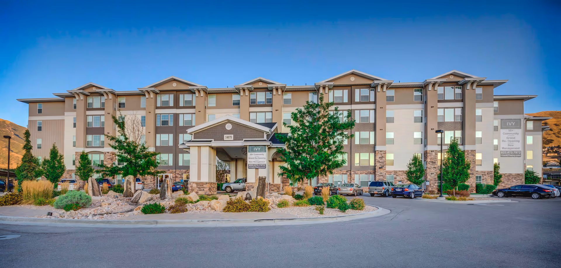 Exterior view of The Ivy at Draper, a multi-story senior living community building with a covered entrance, landscaped rock garden, trees, and parked cars in front under a clear blue sky.