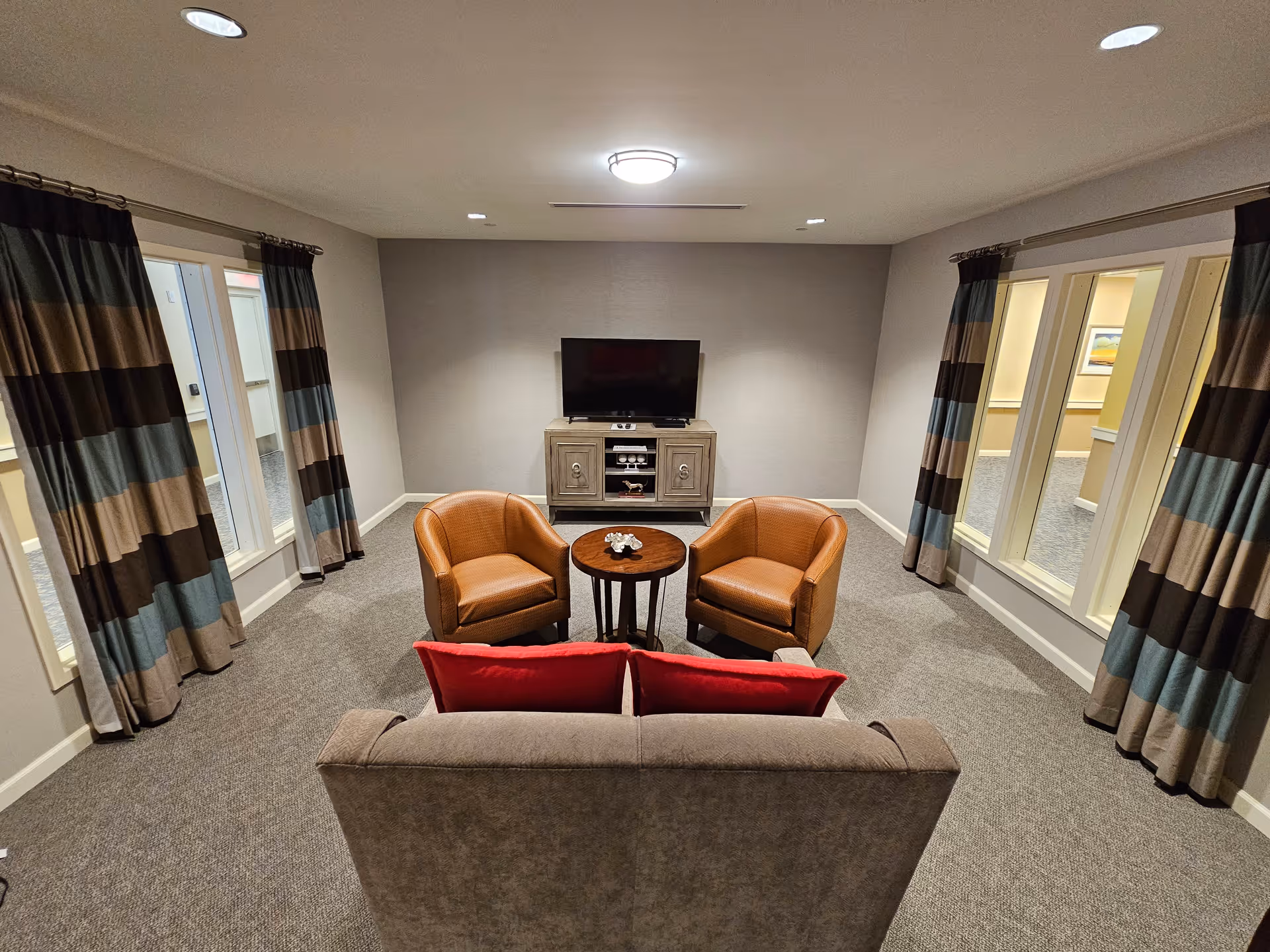 A cozy living room area with a gray sofa featuring two red cushions, two brown leather armchairs, and a small round wooden table in the center. A flat-screen TV is mounted on a cabinet against a plain light gray wall. The room has carpeted flooring and windows on both sides with striped curtains in shades of brown, blue, and beige.