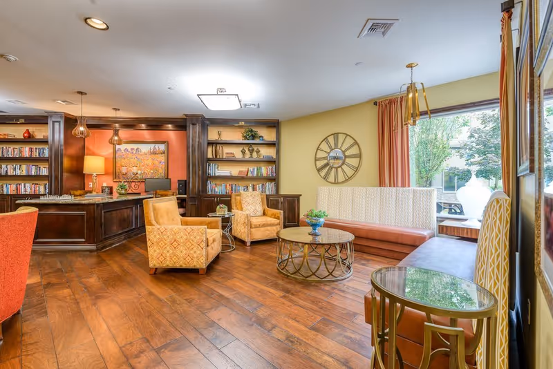 Cozy, well-lit common room with upholstered chairs, a round coffee table, built-in bookshelves and a large window.