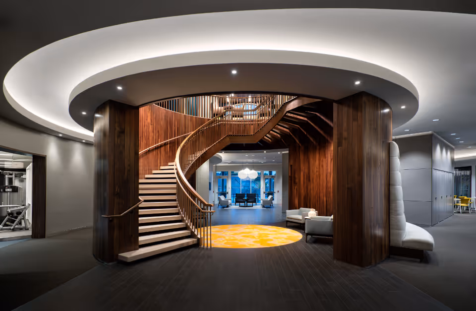 Modern interior space featuring a curved wooden staircase with a circular ceiling light above. The area includes wood-paneled walls, a bright yellow circular rug, and seating areas with armchairs. Adjacent rooms and hallways are visible, including a gym area on the left and a dining or meeting space on the right.