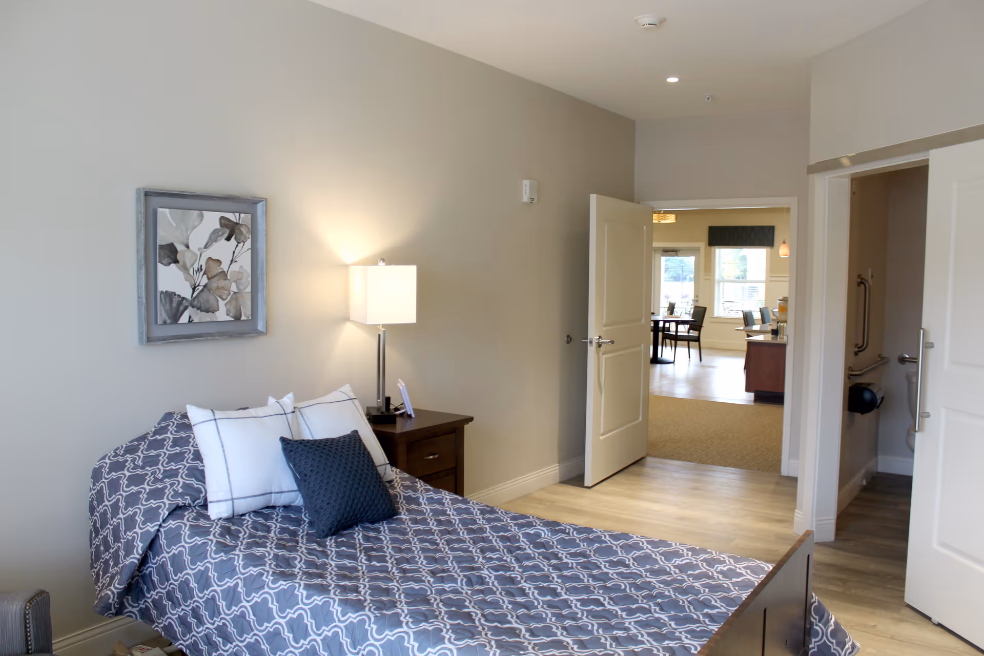 A neatly made single bed with a gray and white patterned bedspread and three pillows in a room with light gray walls. A wooden nightstand with a modern lamp is beside the bed. A framed floral artwork hangs above the bed. The door to the room is open, showing a dining area with tables and chairs in the background. Another door on the right leads to a bathroom with grab bars visible.