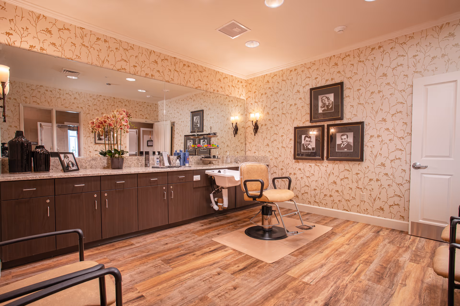 Interior of a salon room with a single beige salon chair in front of a wash basin. The room has wood flooring, floral wallpaper, a large mirror above a long countertop with cabinets, and framed black and white portraits on the wall. There are also decorative vases and a potted orchid on the countertop.