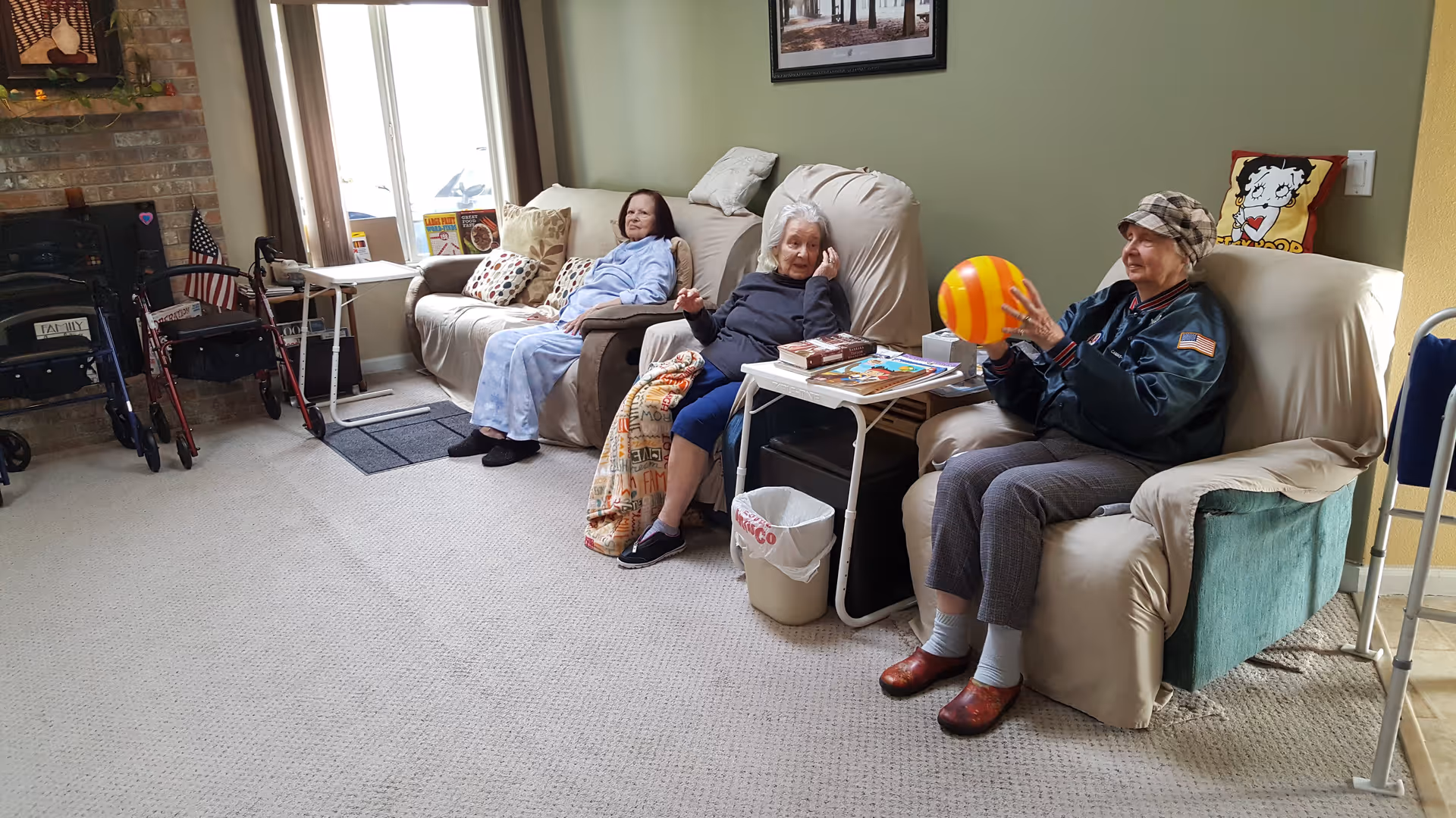 Three elderly women sitting in a living room on comfortable chairs and a sofa. One woman is holding a yellow and orange striped ball, while the others are seated nearby with blankets and books on a small table. The room has a carpeted floor, a brick fireplace, and windows letting in natural light. Walkers are positioned near the fireplace.