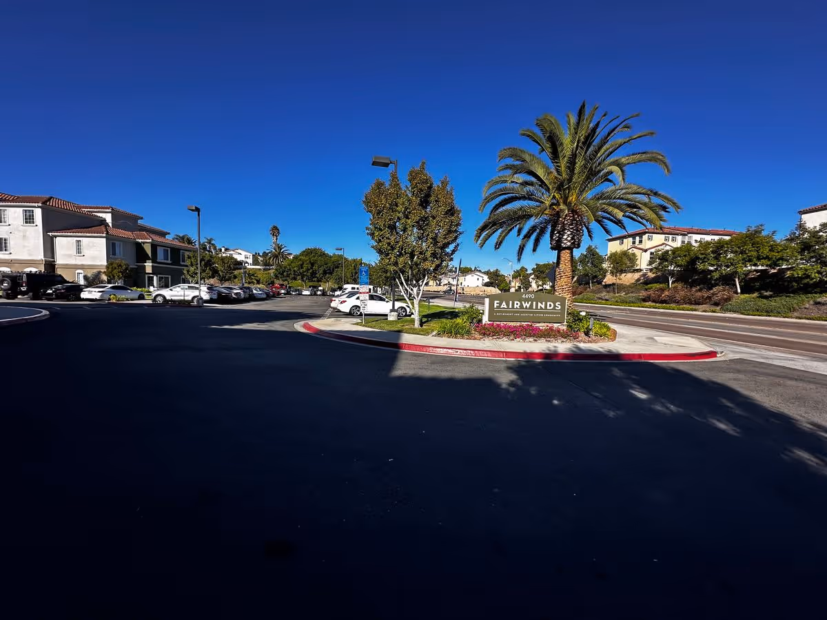 Entrance to the Fairwinds community showing a palm-tree-flanked sign, surrounding buildings, parked cars, and a clear blue sky.