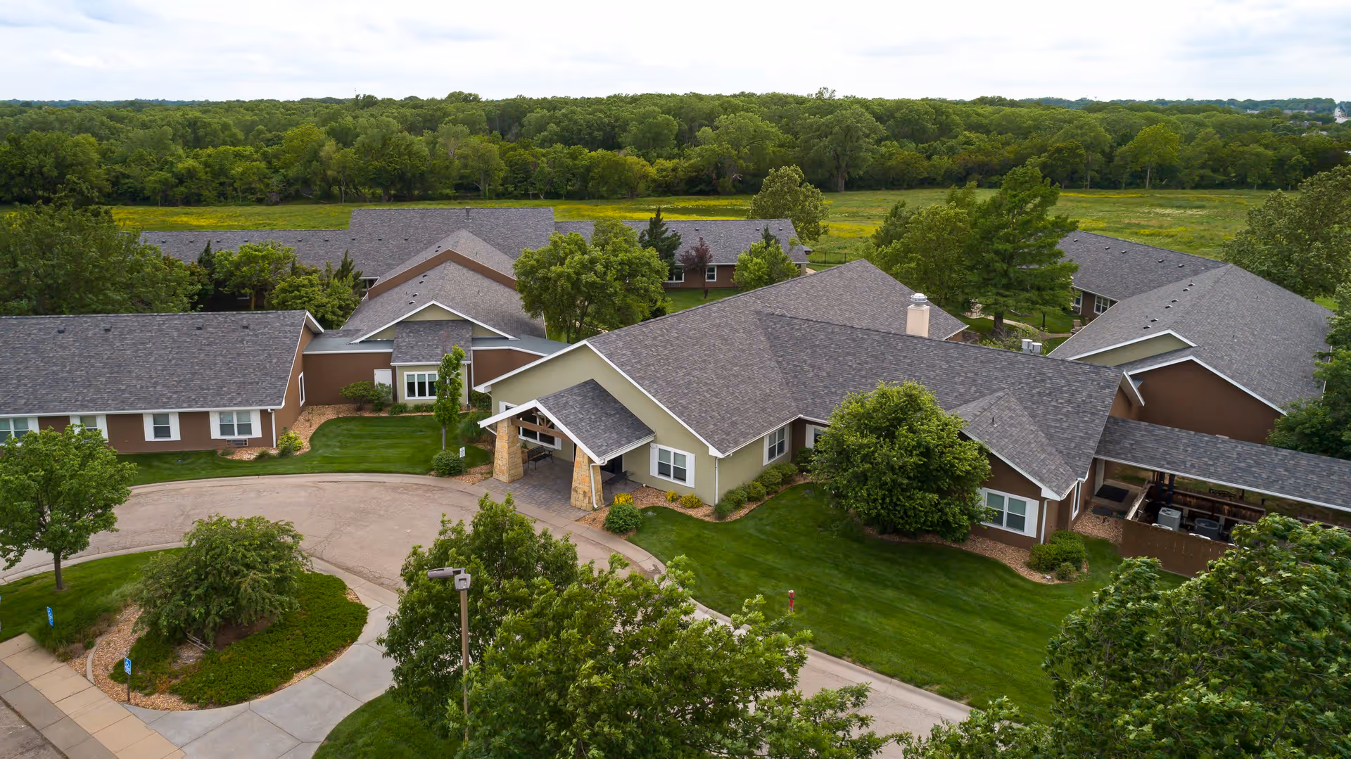 Aerial view of Homestead Assisted Living & Memory Care of Wichita, showing multiple single-story buildings with gray roofs surrounded by green lawns, trees, and landscaping. The facility is set against a backdrop of dense forest and open fields.