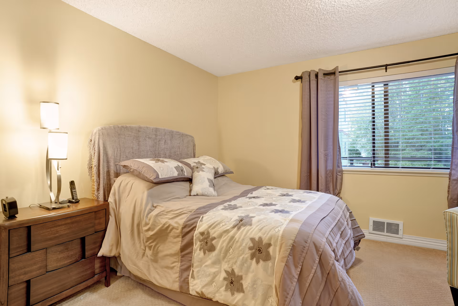 A cozy bedroom with a bed covered in beige and floral-patterned bedding. There is a wooden nightstand with a modern lamp and a phone on it. A window with brown curtains and blinds lets in natural light, and the walls are painted a soft beige color.