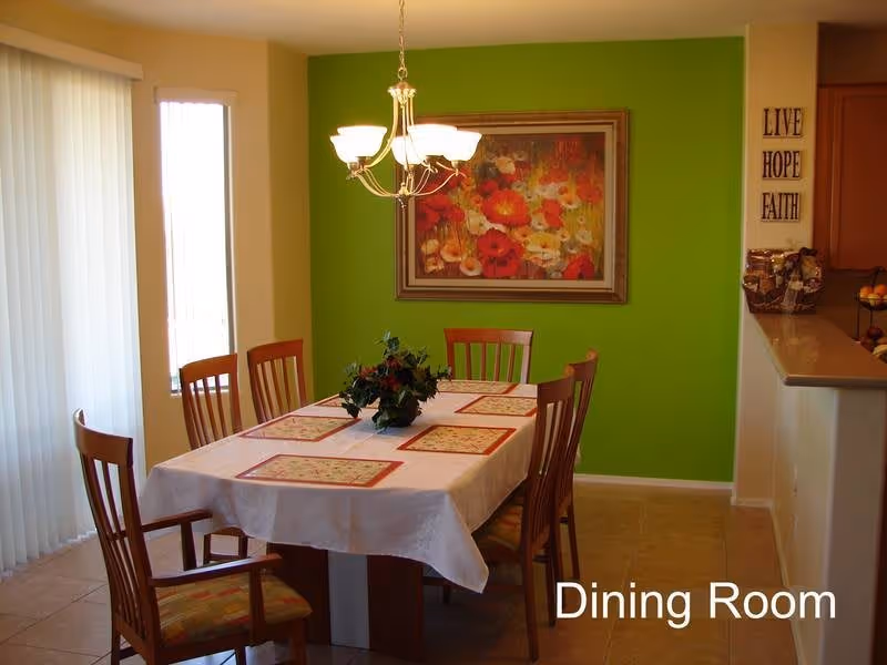 Dining room with a table for six, wooden chairs, chandelier, green accent wall and a floral painting.