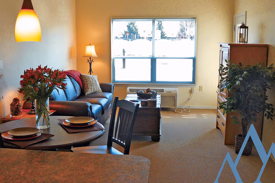 A cozy living room area with a dark leather sofa adorned with a pillow and a red throw blanket. In front of the sofa is a wooden coffee table with decorative items. To the left, there is a small round dining table set with two plates and a vase of red flowers. A floor lamp stands beside the sofa, and a large window with blinds lets in natural light. On the right side, there is a wooden cabinet and a potted plant. The room has beige walls and carpeted flooring.