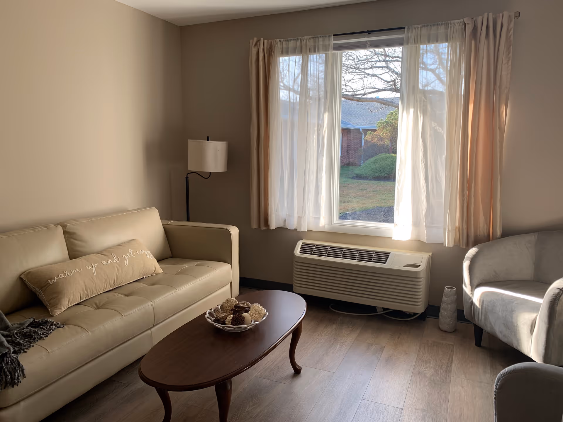 A cozy living room with a beige leather sofa adorned with a decorative pillow that says 'warm up and get cozy', a wooden coffee table with a bowl of decorative items, a floor lamp, a cushioned armchair, and a large window with sheer and beige curtains letting in natural light. Outside the window, some greenery and a brick building are visible.