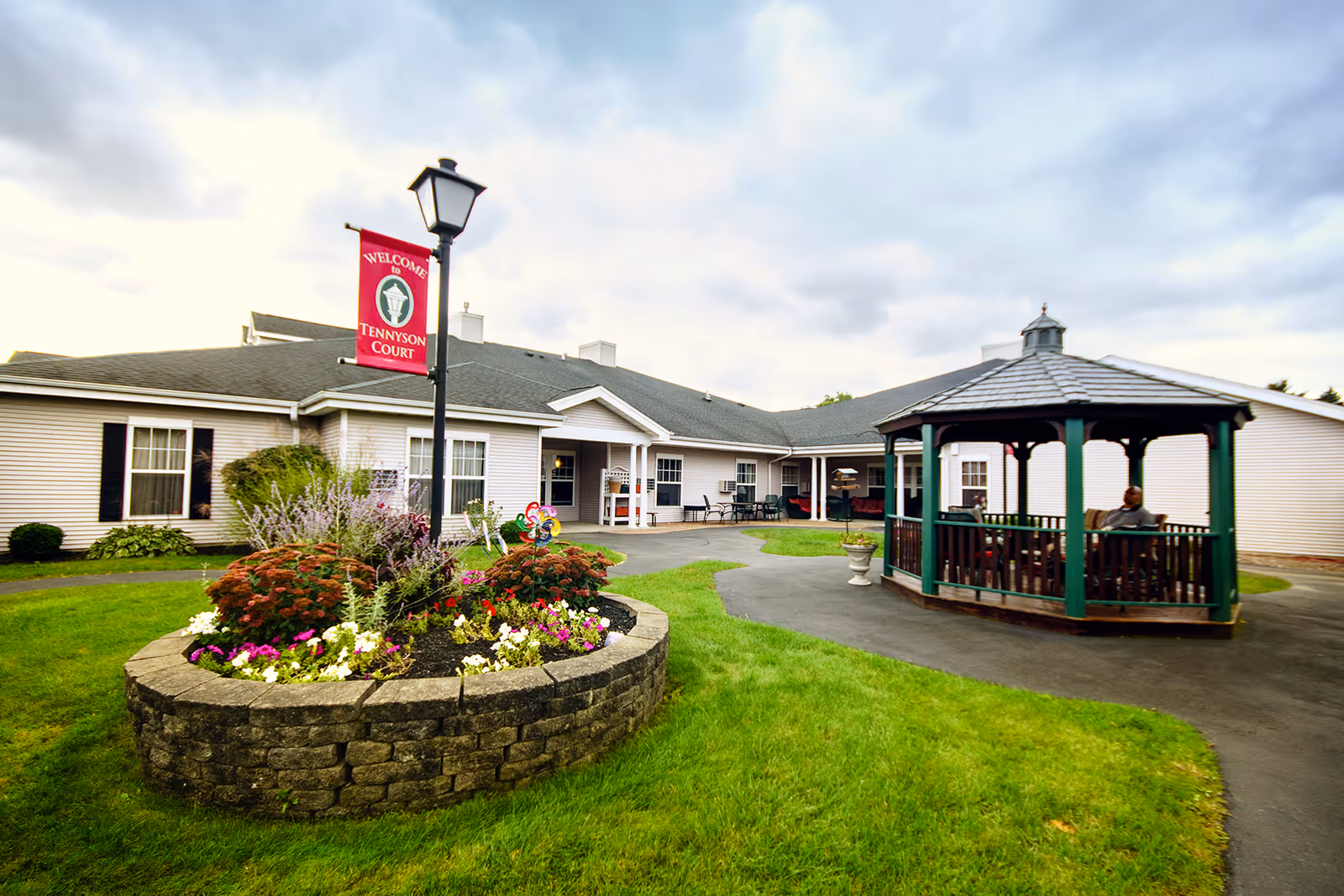 Outdoor view of Tennyson Court senior living facility showing a single-story building with beige siding and black shutters. In the foreground, there is a circular flower bed with colorful flowers and shrubs surrounding a lamp post with a red banner that reads 'Welcome to Tennyson Court.' To the right, there is a wooden gazebo with a person sitting inside, and a paved walkway curves around the garden area.