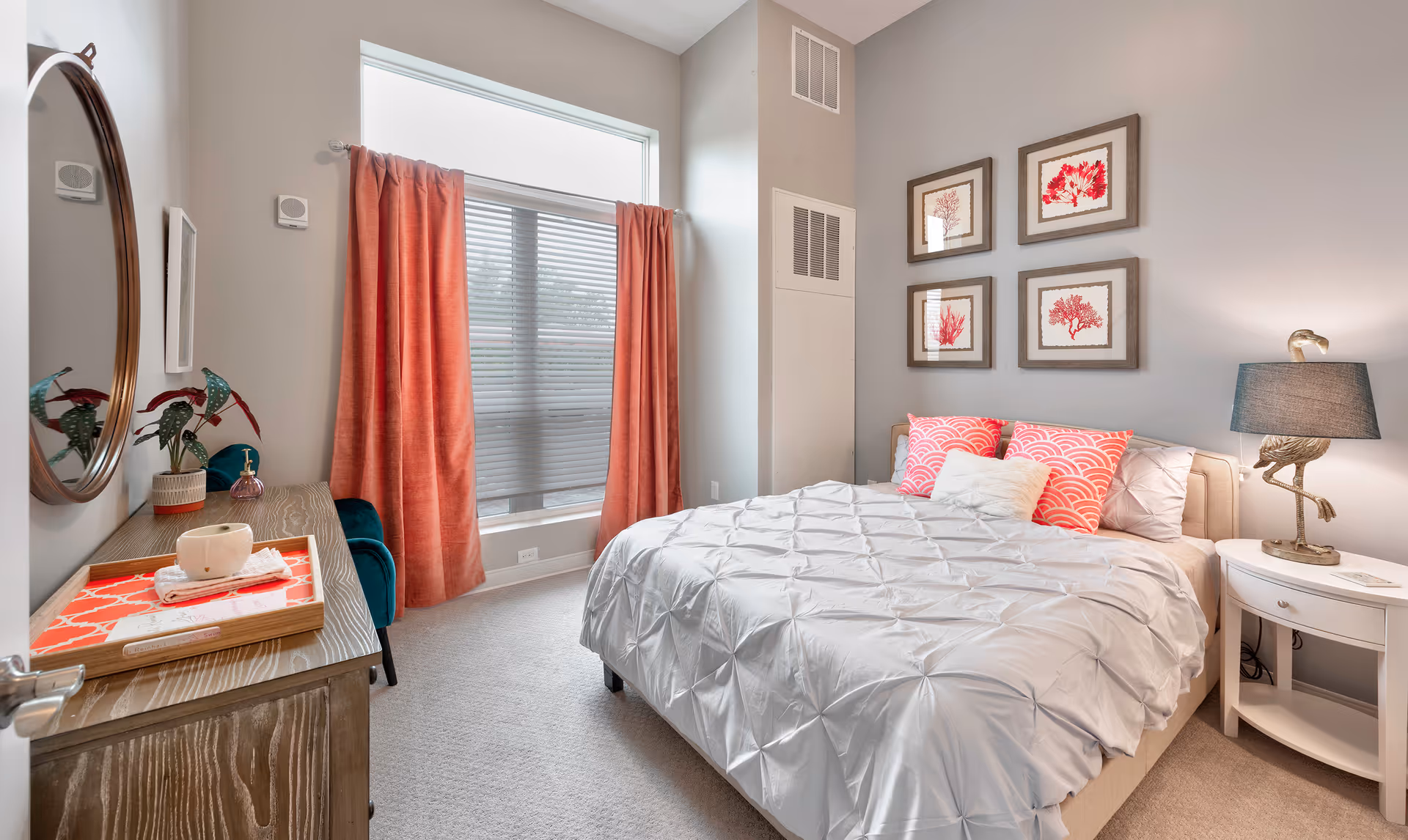 A bright and cozy bedroom with a large window covered by coral-colored curtains and blinds. The bed is neatly made with a white comforter and coral and white decorative pillows. On the right side of the bed is a white nightstand with a lamp featuring a bird-shaped base. Above the bed are four framed coral-themed artworks. On the left side of the room is a wooden dresser with a round mirror above it, a small plant, and a tray with a cup and napkin.