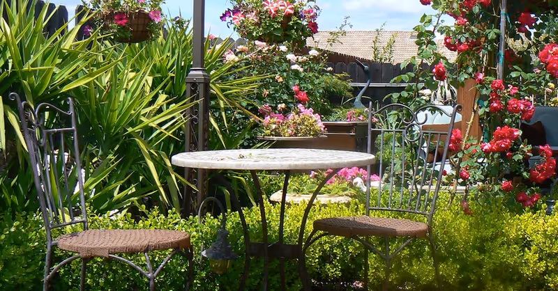 A small outdoor garden seating area with a round metal table and two metal chairs with woven seats. The area is surrounded by lush green plants and vibrant pink and red flowers, with a wooden fence and rooftops visible in the background under a partly cloudy sky.