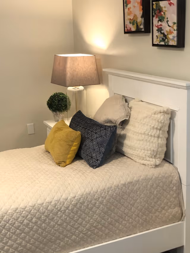 A neatly made bed with a white quilted bedspread and four decorative pillows in yellow, dark blue, and white colors. Next to the bed is a white nightstand with a small green potted plant and a table lamp with a beige shade. Two colorful abstract paintings hang on the wall above the headboard.