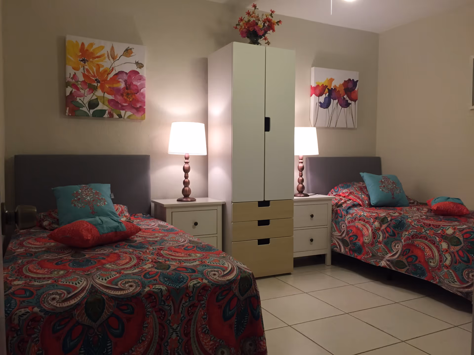 A bedroom with two single beds, each with colorful patterned bedspreads and decorative pillows. Between the beds is a white nightstand with a lamp on each side, and a tall white wardrobe with drawers in the center. Above each bed hangs a floral painting. The room has tiled flooring and beige walls.