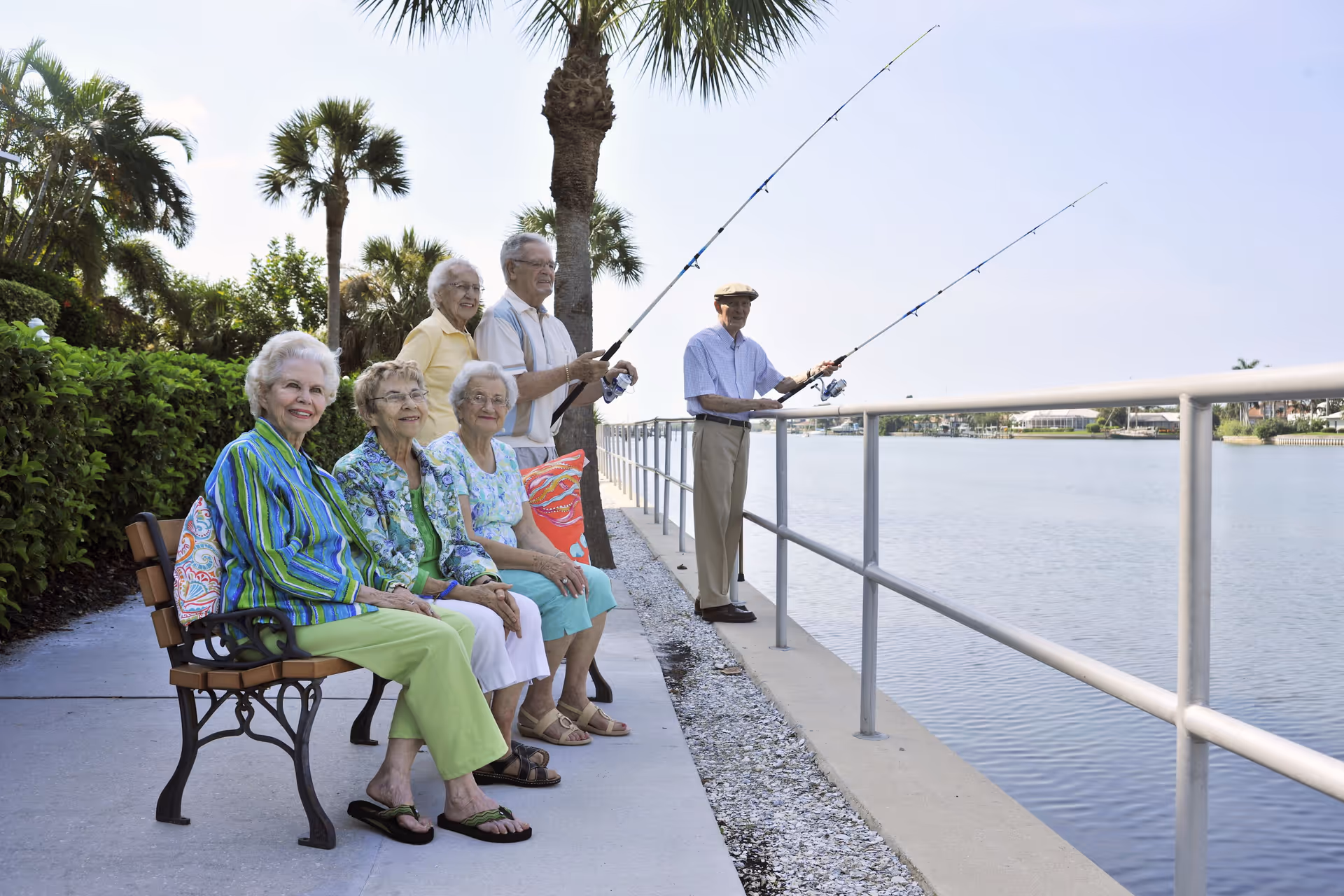 A group of elderly people enjoying a sunny day by a waterfront. Three women are sitting on a bench smiling, while two men and one woman stand behind them fishing with rods. Palm trees and greenery surround the paved walkway beside the water.