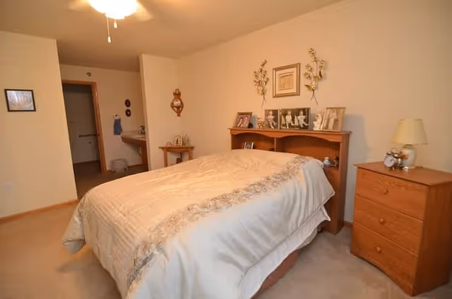 Bedroom with a bed and wooden headboard displaying framed photos, a nightstand and dresser, and an open doorway to a bathroom/vanity area.