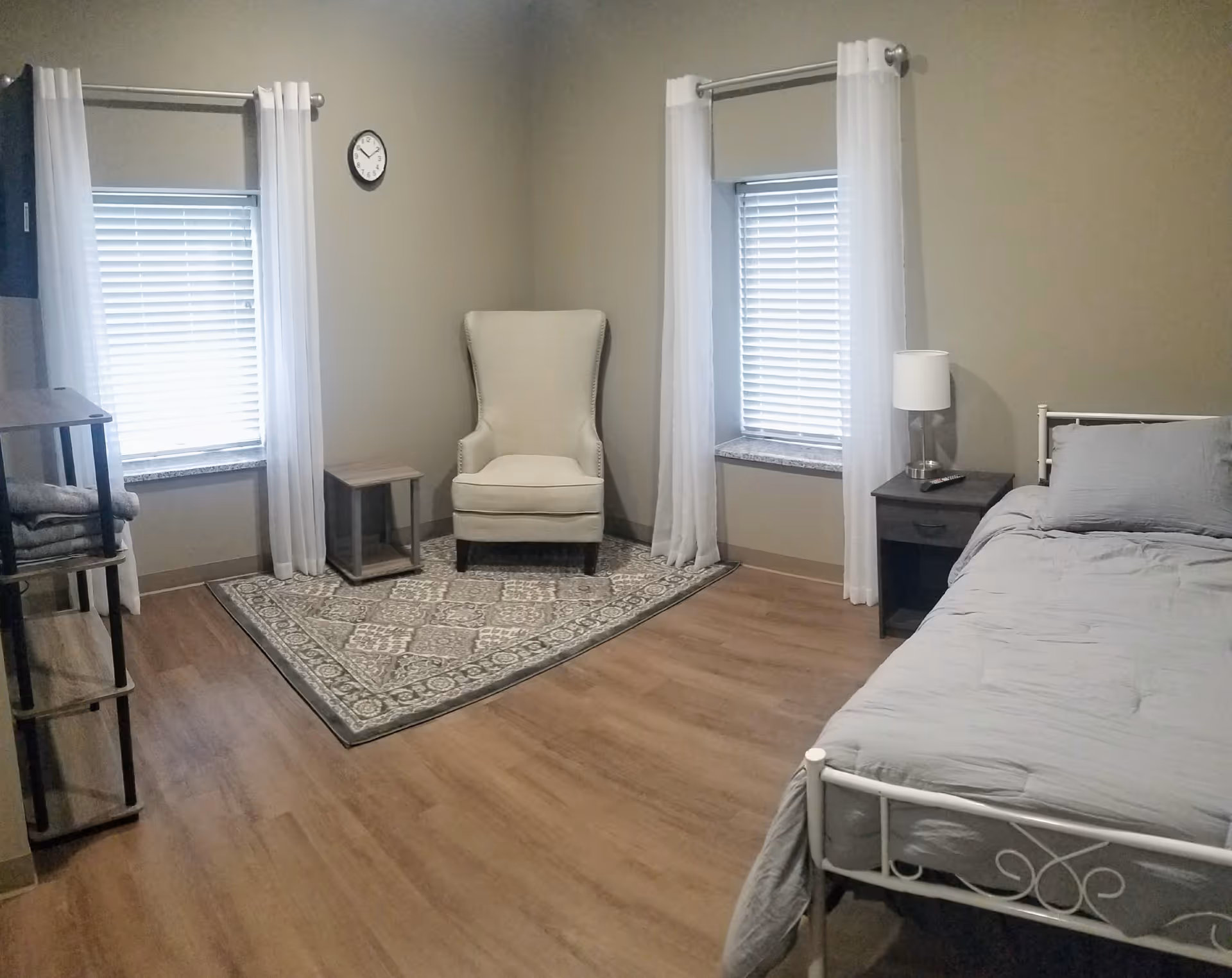 Private bedroom with a single metal bed, armchair on a patterned rug, nightstand with a lamp, and two windows with white curtains.