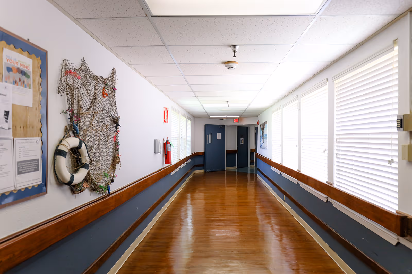 A well-lit hallway in a healthcare facility with wooden handrails on both sides and windows with blinds on the right. The left wall features a bulletin board with notices and a decorative fishing net with a life preserver. The floor is polished wood, and there are double doors at the end of the hallway.