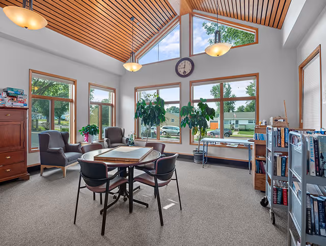 Bright communal lounge with a round table and chairs, armchairs, bookshelves, large windows and plants under a wood-paneled vaulted ceiling.
