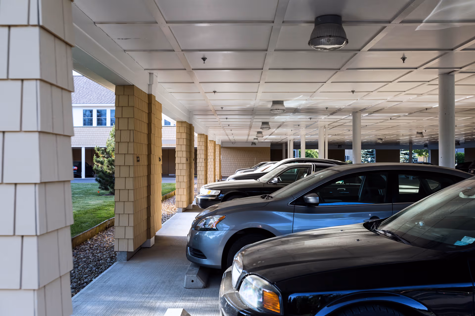 Covered parking area with several parked cars under a white ceiling supported by beige columns. The area is adjacent to a building with beige siding and a small landscaped section with grass and a tree.