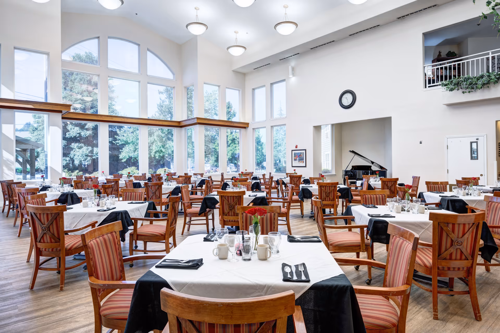 A spacious dining room with multiple wooden tables and chairs arranged neatly. Each table is covered with a white tablecloth and black napkins, set with glasses, cups, and silverware. Large windows with an arched design allow natural light to fill the room, and a black grand piano is visible in a recessed area on the right wall. The room has high ceilings with modern light fixtures and a clock on the wall.