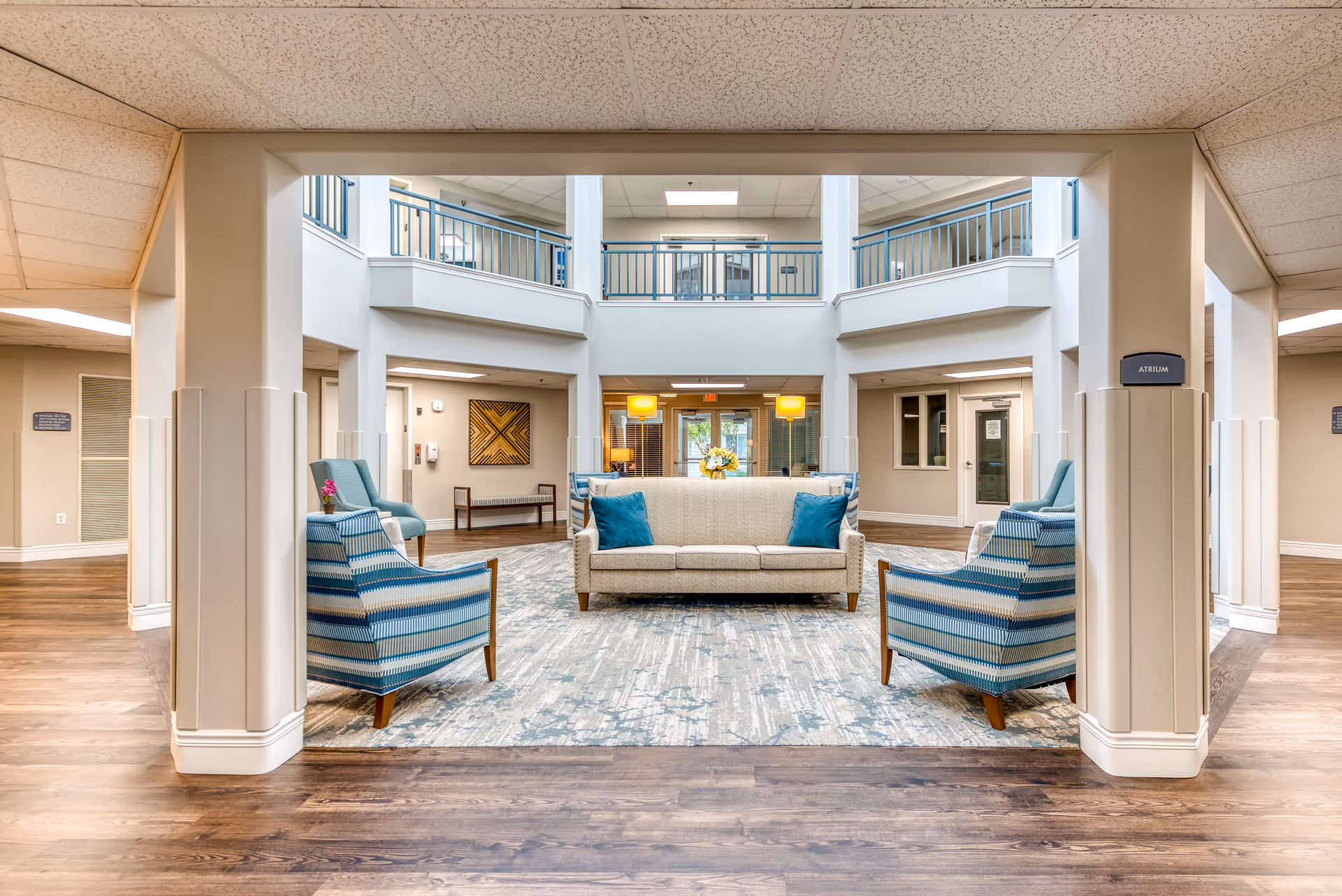 A bright and spacious senior living facility atrium with a beige sofa adorned with blue pillows, two striped blue armchairs, and a patterned area rug on a wooden floor. The atrium features a two-story design with a balcony railing on the upper level, neutral-colored walls, and ceiling lights providing ample illumination.