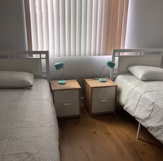 A bedroom with two single beds separated by two wooden nightstands, each with a small green desk lamp. The beds have light-colored bedding and pillows. Behind the beds is a window with vertical blinds partially closed, allowing soft natural light into the room. The floor is wooden.