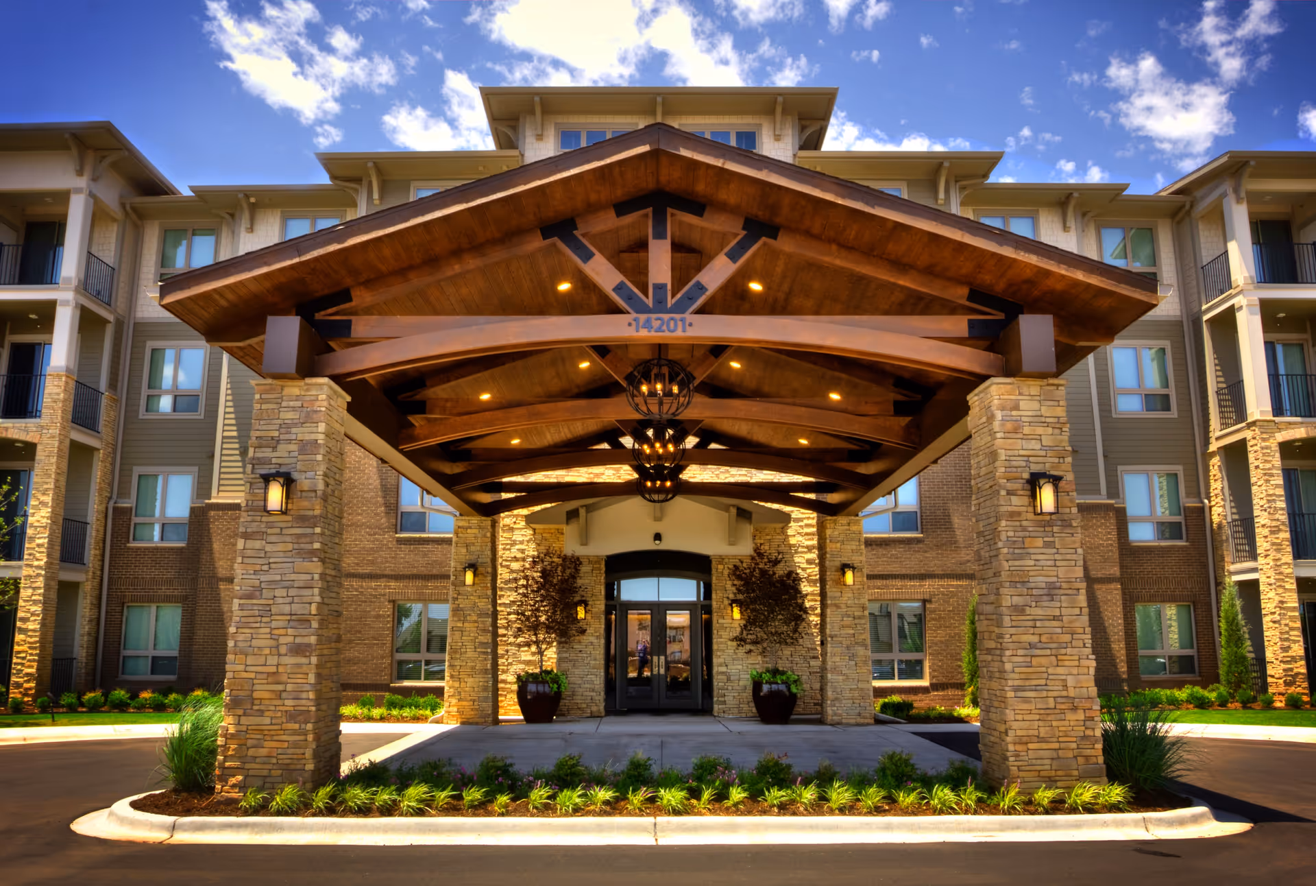 Front entrance of a senior living facility named Album Quail Springs, featuring a large covered driveway with wooden beams and stone pillars, surrounded by a multi-story building with balconies and windows under a partly cloudy blue sky.