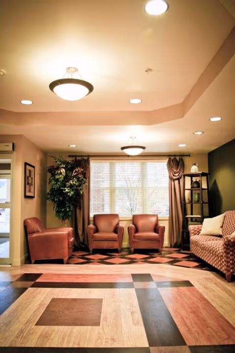 A cozy living room area with three brown leather armchairs and a patterned sofa. The room features a large window with blinds and brown curtains, a tall green plant, a shelving unit with decorative items, and ceiling lights. The floor has a geometric patterned design in shades of brown and beige.
