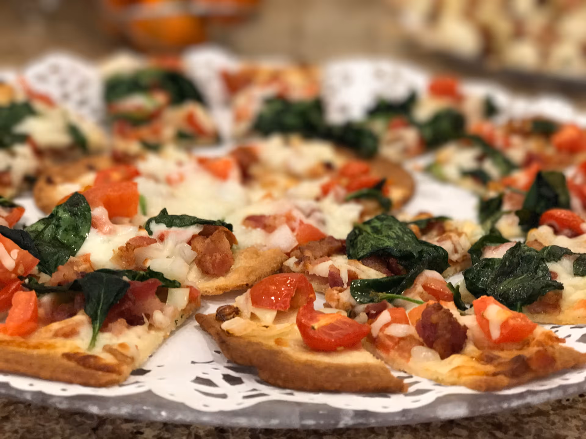 Close-up view of several slices of pizza topped with spinach, diced tomatoes, onions, bacon, and melted cheese on a decorative white doily.