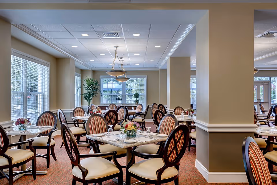 A bright and spacious dining room with multiple tables and chairs arranged neatly. Each table is set with glasses, plates, and floral centerpieces. Large windows with blinds allow natural light to fill the room, and a hanging light fixture is visible from the ceiling. The walls are painted beige, and there are decorative plants near the windows.
