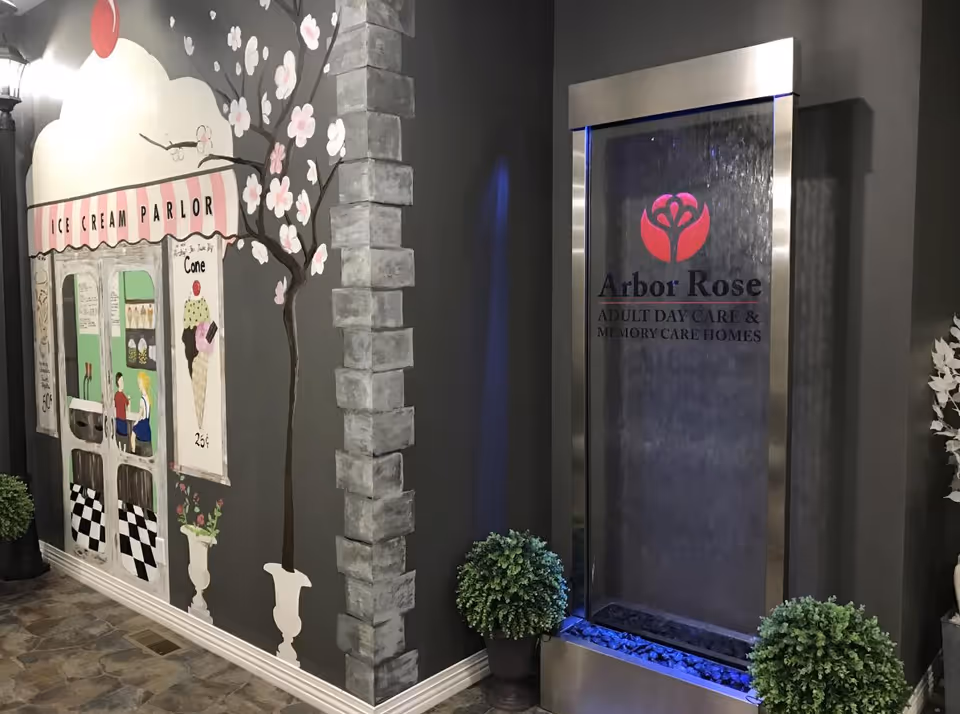 Indoor area with a decorative wall mural of an ice cream parlor featuring a tree with pink blossoms and a sign advertising cones for 25 cents. Next to the mural is a modern water feature with the Arbor Rose logo and text reading 'Arbor Rose Adult Day Care & Memory Care Homes.' Two potted plants are placed on either side of the water feature.