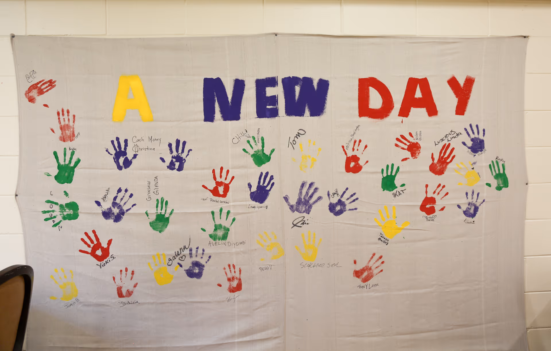 A large white cloth hanging on a wall with colorful handprints in red, green, purple, and yellow. The words 'A NEW DAY' are painted in large letters across the top in yellow, purple, and red. Many handprints have names written next to them.