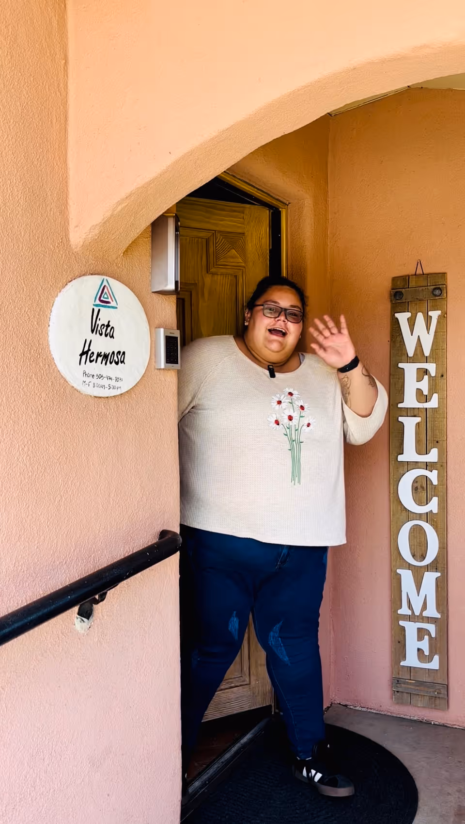 A person wearing glasses waves from the doorway of a peach-colored entryway beside a round 'Vista Hermosa' sign and a vertical 'WELCOME' sign.
